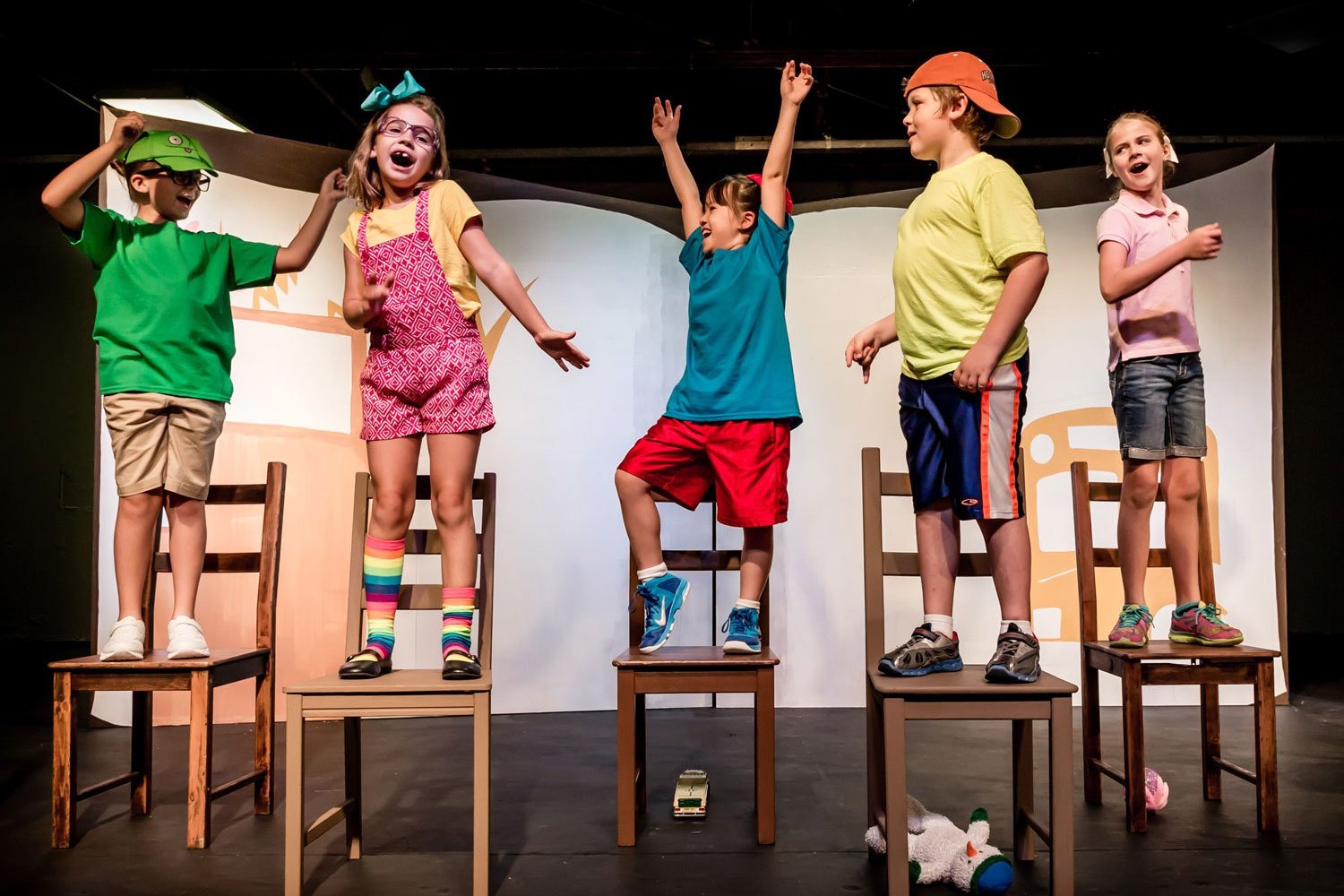 Five kids standing on wooden chairs mid-scene, arms up and laughing, during an improv game